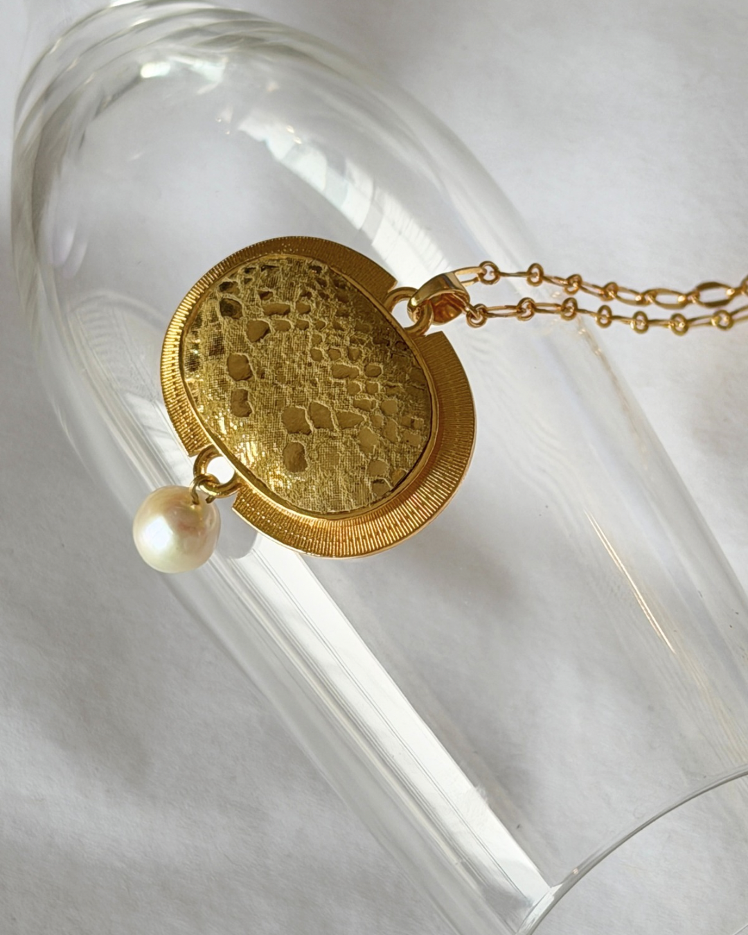 A gold pendant necklace with a textured, foiled gold leather on an oval face and a dangling white pearl, displayed on a clear glass surface.