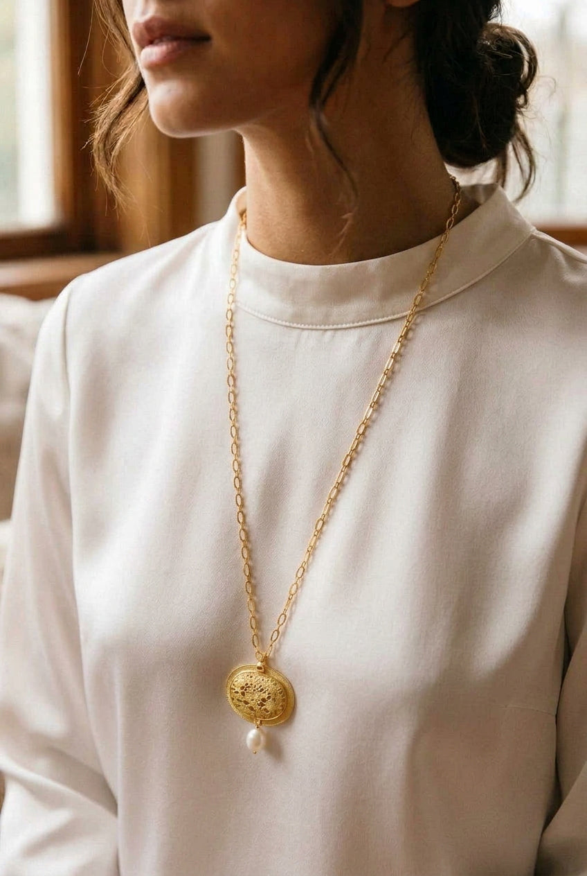 A gold chain necklace with a round textured gold pendant and a small white pearl drop, laid flat on a white fabric surface.