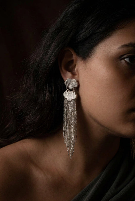 A close-up side profile of a woman wearing a long, textured silver dangle earring with cascading fringe beads.