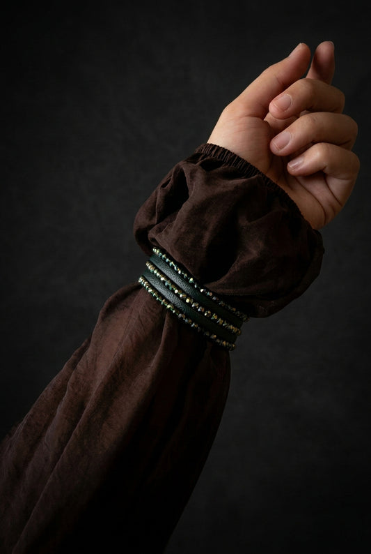 A close-up of a person's hand and forearm wearing a dark brown sheer puff sleeve with a decorative beaded cuff, set in a warmly lit living room interior.