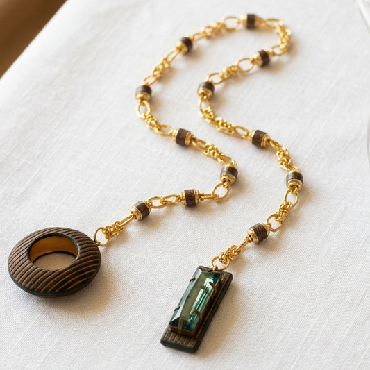 A gold chain necklace with dark brown wooden beads and two wooden pendant charms, one circular with a striped pattern and one rectangular with a green inlay, displayed on a white textured fabric.